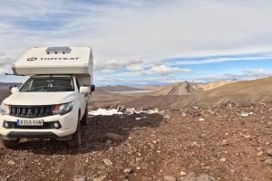 Pickup camper TomyKat en ruta de overlanding en la cima de una montaña con lago y pico nevado al fondo